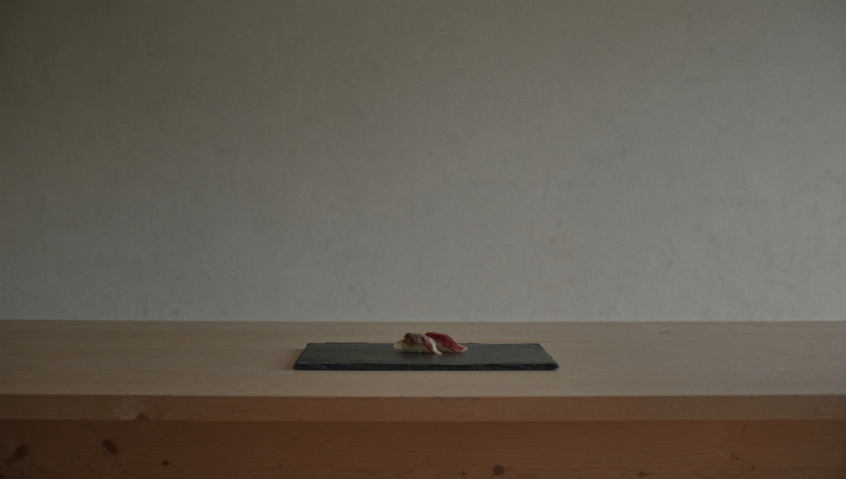 Two pieces of nigiri on a dark slate plate, set on a pale hinoki counter in soft window light